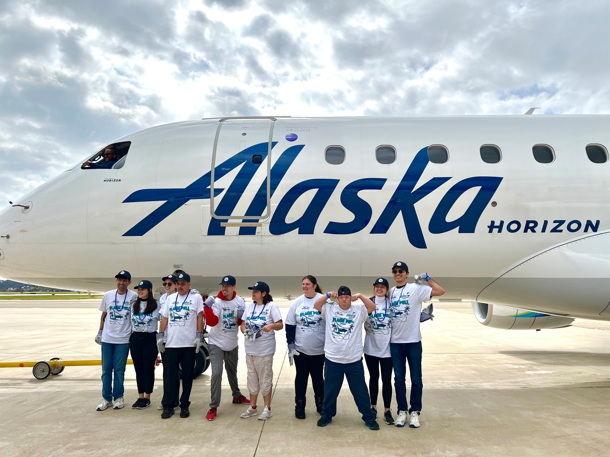 A group of people stand in front of an Alaska Airlines plane wearing Plane Pull t-shirts